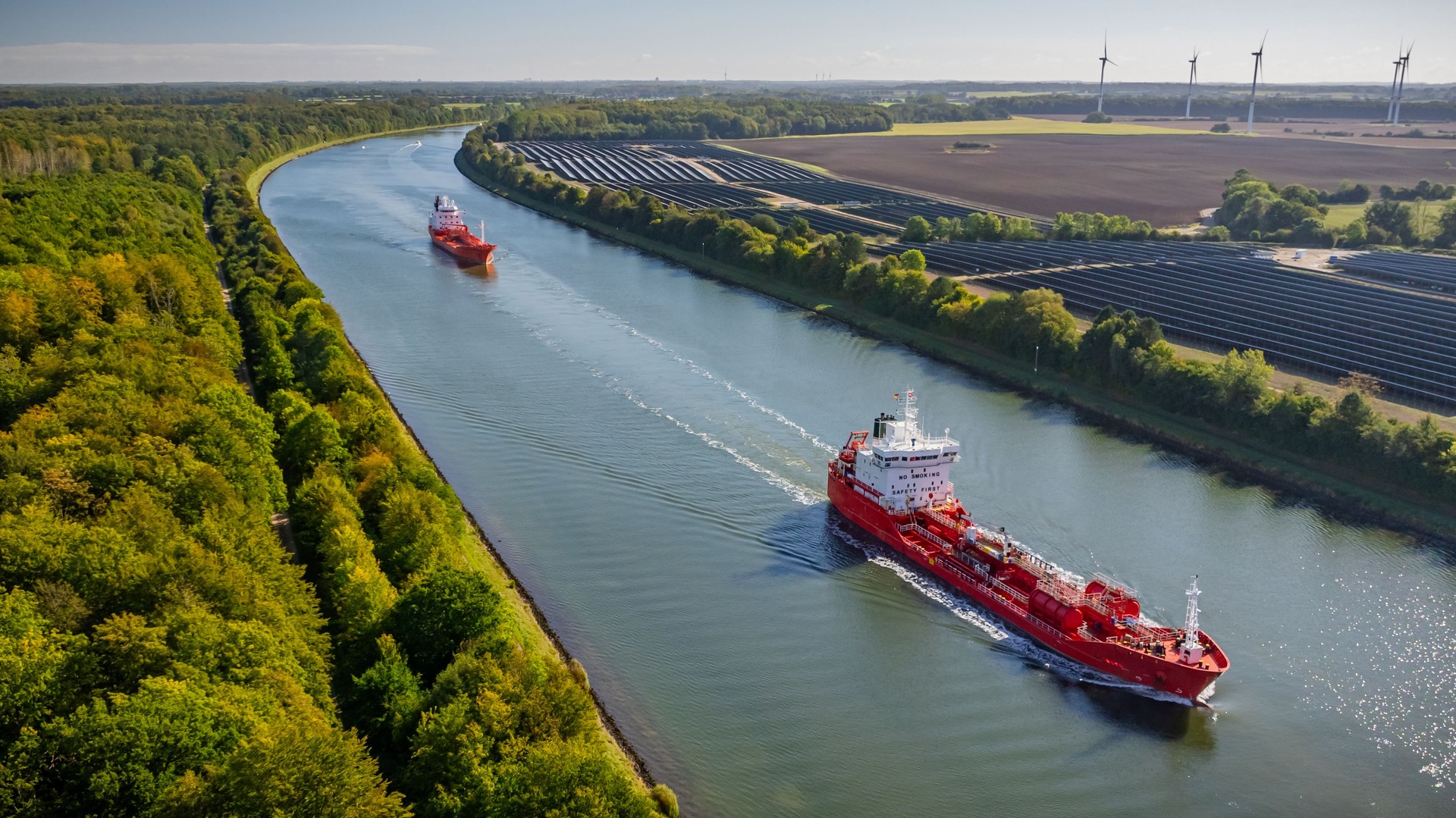 Tanker transporting fossil fuels along waterway, sailing past canal bordered by solar farm. Traditional and modern energy. Concept of transition from fossil fuels to renewable energy.